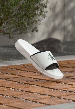 Witte Calvin Klein slippers met zwarte logo-strap, gepositioneerd boven houten tegels en kleine grijze kiezelstenen buiten.