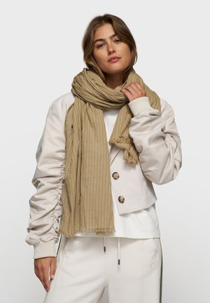 Sciarpa beige ammuffita con bordi frangiati, avvolta attorno al collo. Tessuto morbido, leggero, che completa una giacca e pantaloni di colore chiaro.