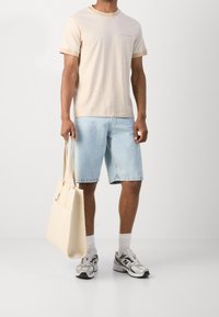 Lysebeige T-shirt med korte ærmer, lyseblå denimshorts, grå sneakers og en beige indkøbs taske. Outfit kombinerer afslappede stoffer og farver.