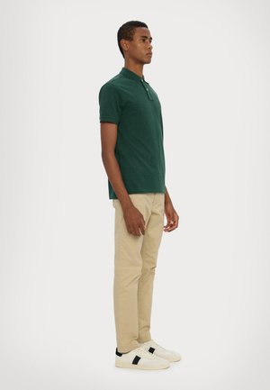 Žalias polo marškinėlis trumpomis rankovėmis, plokščiu apykakle ir mygtukų eiliu. Suderintas su kreminės spalvos kelnėmis ir baltais sportbačiais su juodais akcentais.