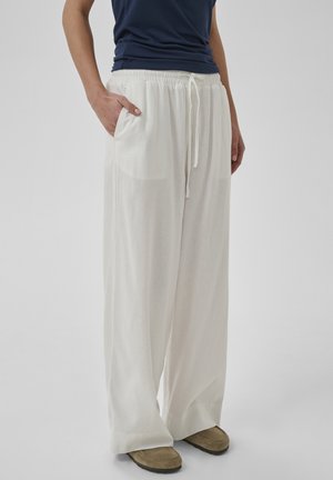 Persoon draagt witte wijde broek met trekkoord in de taille, donkerblauw topje en beige schoenen, staat met één hand in de zak.