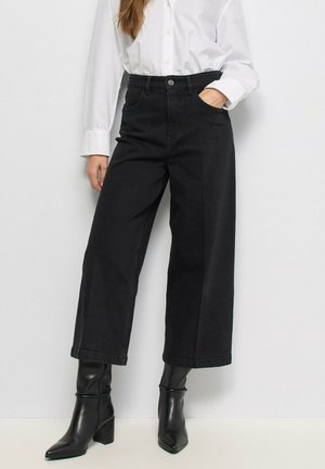 Mujer con vaqueros negros cortos de pierna ancha, camisa blanca abotonada y botines negros de tacón con punta afilada.