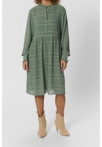 Abito verde con colletto rotondo, maniche lunghe, vita arricciata e sottili righe orizzontali. Abbinato a stivali alla caviglia beige.