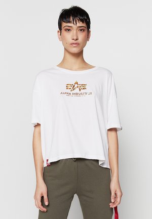 Person mit kurzen dunklen Haaren, die ein weißes Alpha Industries T-Shirt und olivgrüne Hose trägt, steht vor einem schlichten hellen Hintergrund.