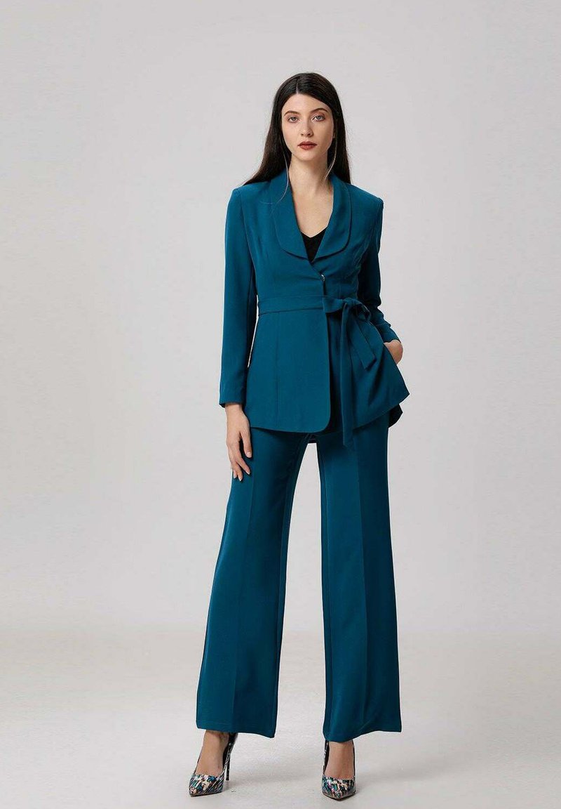 Femme en tailleur teal avec ceinture et haut noir, portant des talons hauts à motifs, se tenant devant un fond clair.