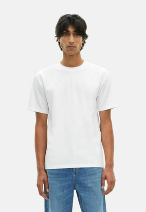 Junger Mann mit mittellangen dunklen Haaren, der ein schlichtes weißes T-Shirt mit kurzen Ärmeln und blaue Jeans trägt, stehend vor einem hellen Hintergrund.