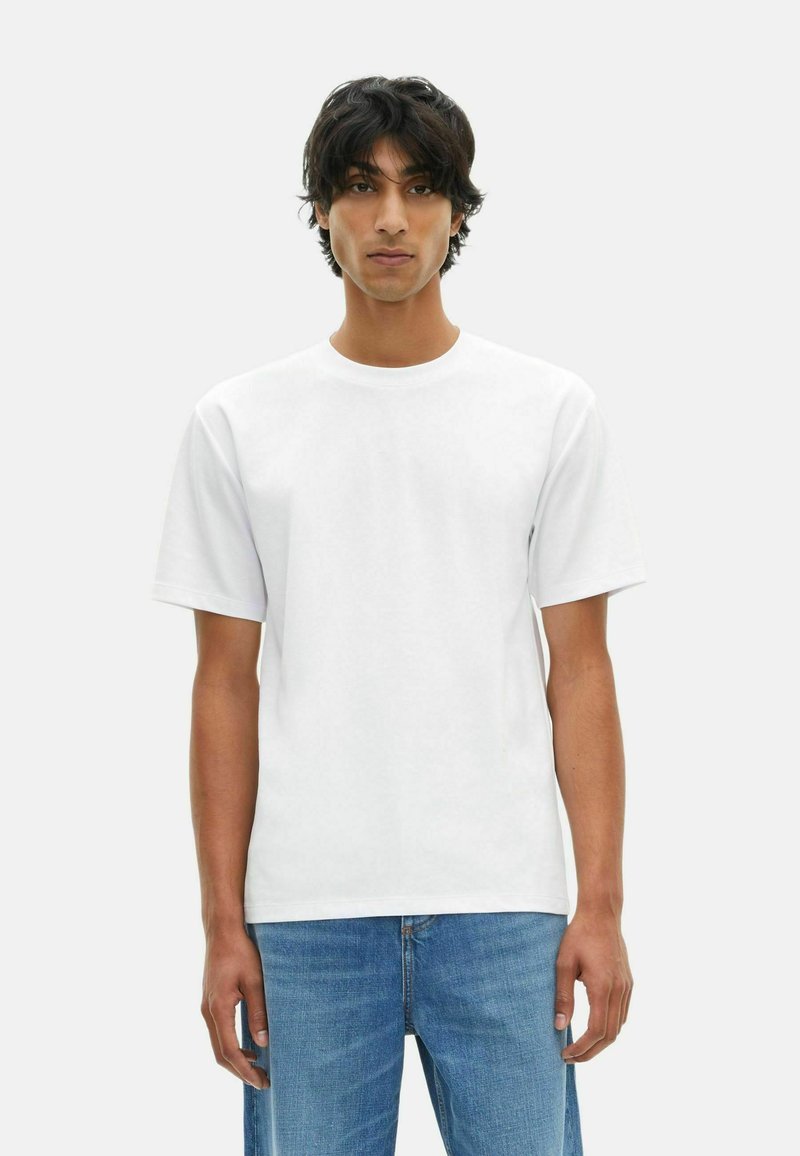 Giovane uomo con capelli scuri di lunghezza media, che indossa una semplice t-shirt bianca a maniche corte e jeans blu, in piedi su uno sfondo chiaro.
