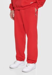 Røde sweatpants lavet af blødt stof, med en afslappet pasform, elastiske manchetter, sidelommer med lynlås og diskret branding på benet. Båret med hvide sneakers.
