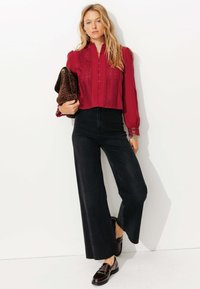 Blouse rouge texturée avec un devant à boutons et des accents en dentelle, associée à un jean noir à taille haute et jambes larges, et des mocassins foncés.