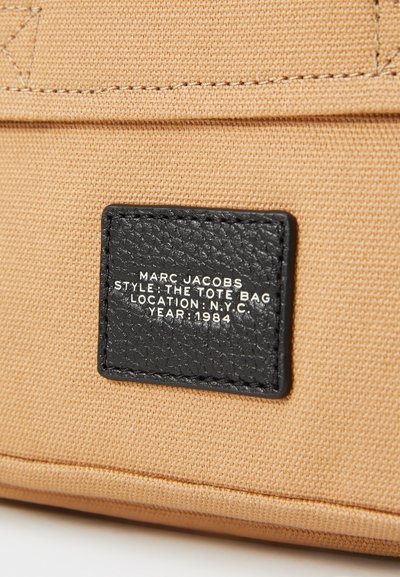 Geantă de tote din țesătură bej, cu un eticheta din piele neagră cu text embosat: "MARC JACOBS," "STIL: GEANTA TOTE," și "AN: 1984."