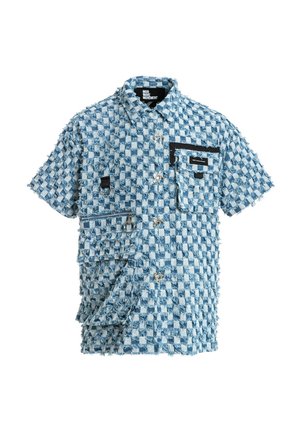 Joven de pie con camisa de manga corta de cuadros azul y blanca y pantalones cortos a juego, calcetines blancos y zapatos beige texturizados sin cordones.