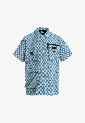 Joven de pie con camisa de manga corta de cuadros azul y blanca y pantalones cortos a juego, calcetines blancos y zapatos beige texturizados sin cordones.