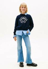 Jersey azul marino con bordados florales blancos y logotipo, superpuesto a una camisa a rayas azul claro. Vaqueros acampanados azules y zapatos de plataforma negros.