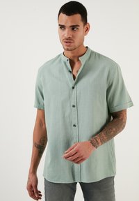 La camisa de manga corta en verde menta presenta un cuello mao, una botonera y una tela texturizada. El modelo lleva jeans grises y muestra tatuajes.