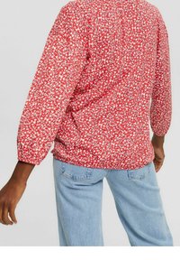 Blusa rossa floreale con maniche lunghe e a sbuffo, caratterizzata da una vestibilità ampia e orlo arricciato. Abbinata a jeans azzurri per un contrasto.