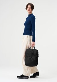 Schwarzer Rucksack mit zwei oberen Griffen, Reißverschlussfach und dekorativen Riemen. Textilstoff mit glatter Oberfläche, minimalistisches Design.