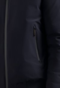 Donker marineblauwe jas met een gladde, matte textuur, een ritszak opzij en contrasterende naden langs de rits en manchetten.