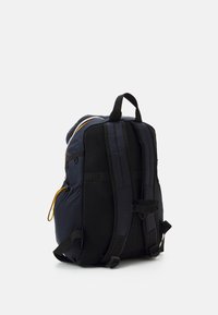 Marineblauer Rucksack mit schwarzen Mesh-Akzenten, gepolsterten Trägern, verstellbaren Schnallen und einer Seiten Tasche mit gelbem Kordelzug.