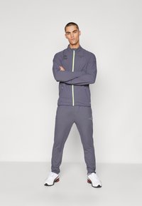 Grijze sportieve trainingsoutfit met een ritsjack voorzien van neon groene accenten en logo-details, gecombineerd met bijpassende taps toelopende broek en witte sneakers.