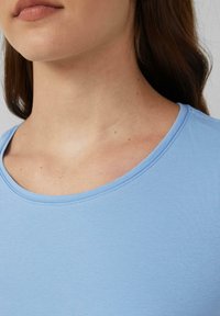 Close-up van het onder gezicht van een vrouw, hals en bovenste borst, die een lichtblauwe shirt draagt met zichtbaar lang bruin haar.