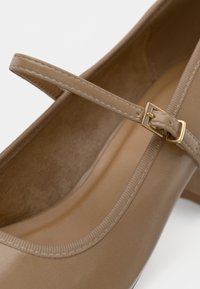 Bežové lakované baleríny typu Mary Jane s tenkým remienkom a malou zlatou prackou na bielom pozadí.