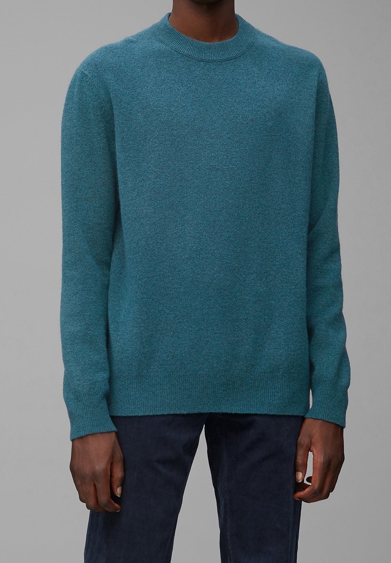 Tealová vlna svetru s kulatým výstřihem, dlouhými rukávy a žebrovanými manžetami. Měkká textura, volný střih a minimalistický design.