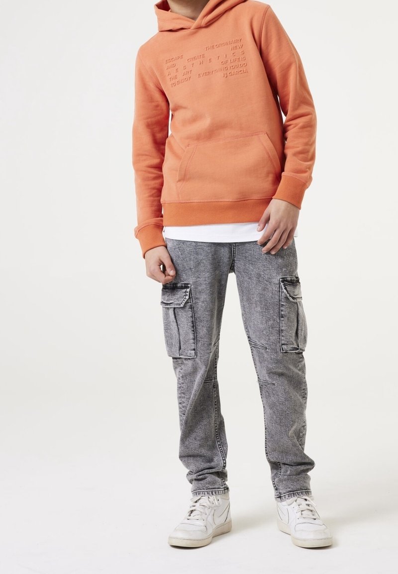 Junge trägt einen orangefarbenen Kapuzenpullover mit erhabenem Text, graue Cargo-Hosen und weiße Sneaker und steht vor einem schlichten weißen Hintergrund.