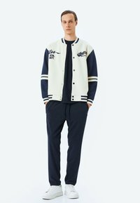 Chaqueta varsity en azul marino y crema con texto atlético y acentos de rayas blancas. Combinada con pantalones oscuros en azul marino y zapatillas blancas.