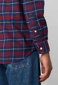 Gros plan sur la main d'une personne à côté d'elle, portant une chemise à carreaux bleu et rouge à manches longues et un jean en denim bleu.