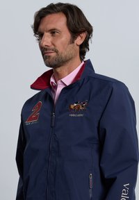 Chaqueta azul marino con un cuello burdeos, que presenta un diseño de caballo bordado y el número "2". Incluye bolsillos laterales con cremallera.