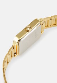 Montre en métal doré avec un cadran rectangulaire, une lunette polie et un bracelet en maillons dorés texturés. Dispose d'une couronne sur le côté droit.