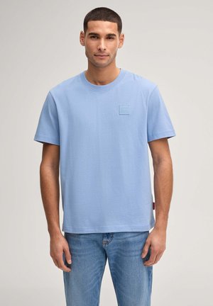 Junger Mann mit kurzen Haaren, der ein hellblaues T-Shirt und blaue Jeans trägt, vor einem schlichten hellen Hintergrund stehend.