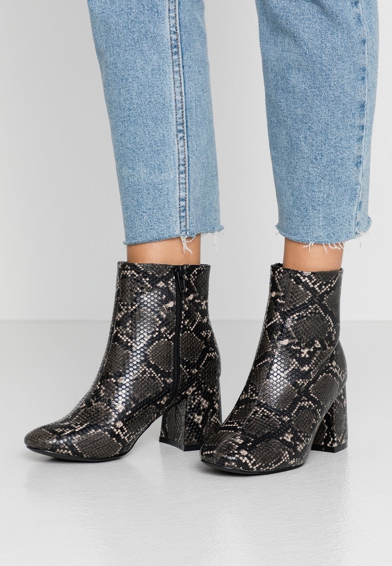 Bottines à imprimé serpent noir et beige avec une texture brillante, talons hauts à bloc et fermetures éclair sur les côtés. Associées à des jeans bleus raccourcis.