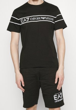 Mand iført sort EA7 Emporio Armani t-shirt med hvide striber og matchende sorte shorts med EA7-logo på venstre ben.