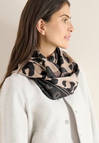 Šátek s leopardím potiskem v barvách pískově hnědé a černé, s jemnou texturou a jednobarevnou černou hranou nesoucí název značky světle zbarveným písmem.