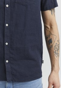 Camisa de manga corta en color navy, fabricada en un material ligero, con un cierre frontal de botones y un bolsillo en el pecho. Superficie texturizada con una etiqueta lateral.