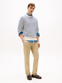 Gestreifter Rundhals-Pullover in Weiß und Marineblau, über einem hellblauen Jeanshemd getragen. Beigefarbene Hose und dunkle Slipper runden das Outfit ab.