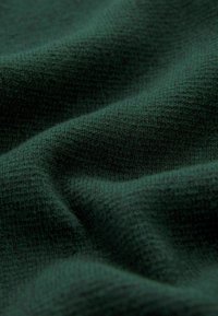 Tessuto verde scuro con una texture che presenta un motivo a maglia morbido, evidenziando effetti di luce e ombra dalle pieghe e dai ripiegamenti.