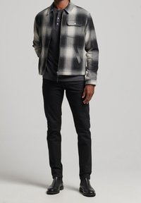 Overshirt-Jacke aus schwarz-weiß kariertem Wollmischgewebe mit einer Vorderseite-Tasche, Reißverschluss und schwarze Slim-Fit-Hose mit Knöchelstiefeln.