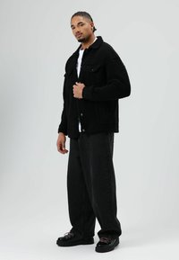 Zwarte sherpa-jas met knoopsluiting, twee borstzakken en een iets croppende pasvorm, gecombineerd met zwarte wijde-jeans en chunky zwarte schoenen.