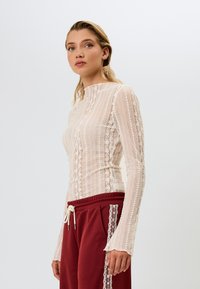 Langermet blonde topp i off-white med høyhalset, med et teksturert mønster. Sammen med burgunder joggebukser med blonder detaljer.