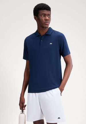 Uomo che indossa una polo blu navy e pantaloncini bianchi, tiene in mano una bottiglia d'acqua bianca e guarda di lato su uno sfondo semplice.