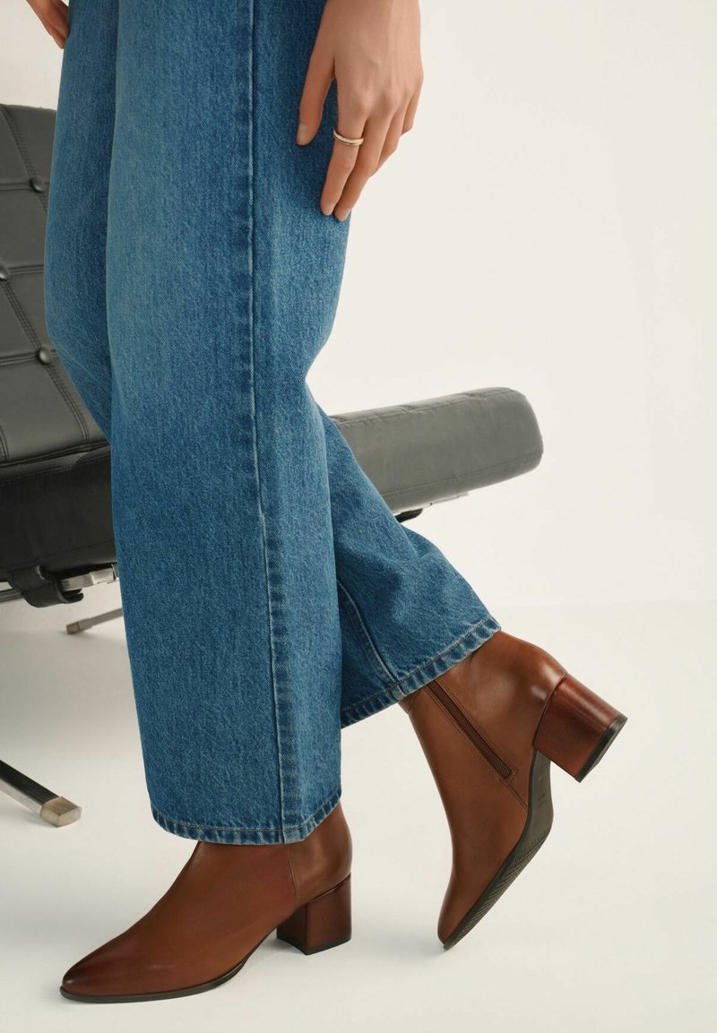 Brązowy skórzany but za kostkę z ostrym noskiem i blokowym obcasem, zestawiony z niebieskimi dżinsami. But posiada boczny zamek błyskawiczny, co ułatwia zakładanie.