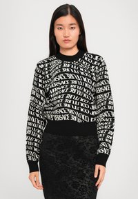 Melns džemperis ar baltu Versace logo dažādās orientācijās. Ribotās piedurknes un apakšmalas, apaļa kakla izgriezums, mīksta struktūra. Saskaņots ar melnu rakstainu svārku.