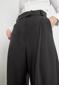 Schwarze weite Stoffhose aus glattem Material. Mit hoher Taille, Gürtelschlaufen und Vorderfalten. Die Taschen sind auf beiden Seiten sichtbar.