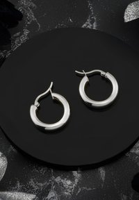 Silverfärgade hoopsörhängen med polerad yta och sömlös design, utrustade med en säker låsning, visade på en cirkulär svart yta.