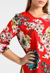 Květinové šaty z červené látky s velkými růžovými a žlutými květinami a zelenými akcenty. Design zahrnuje tříčtvrteční rukávy a kulatý výstřih.