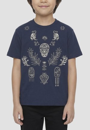 Enfant portant un t-shirt bleu marine avec des motifs blancs complexes comprenant un crâne de sucre, des oiseaux, des plantes, des étoiles et des motifs animaliers.