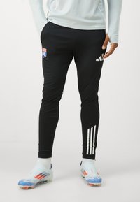 Pantalones deportivos negros con rayas blancas de Adidas, con un parche de logo en el muslo y un diseño ajustado, adecuados para actividades deportivas.