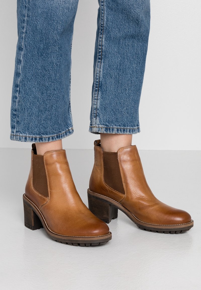 Vrouw die bruine leren enkellaarsjes met hak draagt, gecombineerd met blauwe denim jeans die net boven de enkels zijn afgeknipt, op een lichte vloer.